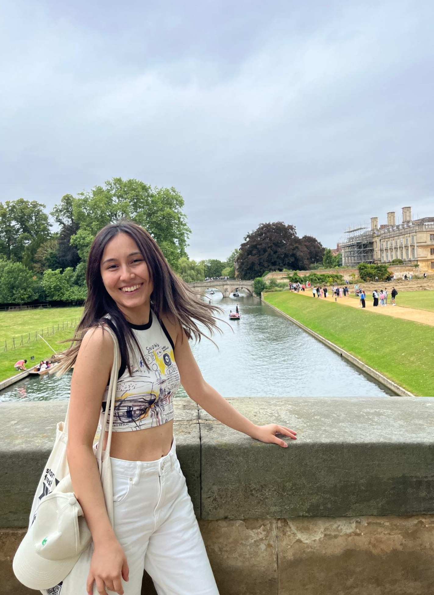 Arzu smiling on a bridge over a river in Cambridge, with punts and a historic building behind her